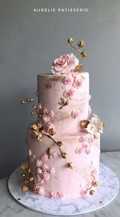 a three tiered cake with pink flowers and gold leaves on the top, sitting on a marble platter