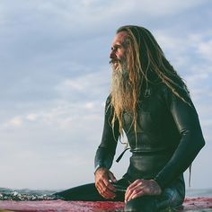 a man with long hair sitting on top of a surfboard next to the ocean