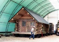 a man walking in front of a log cabin under a large green cover overhang