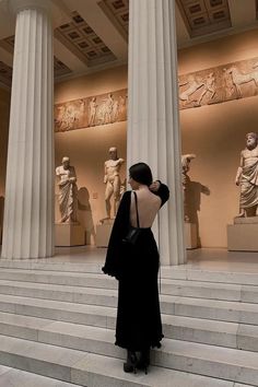 a woman in black dress standing on steps next to white pillars with statues behind her