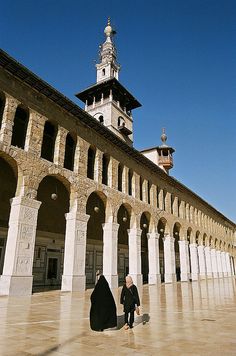 a person in a black outfit is standing near a building with arches and pillars, looking up at the sky