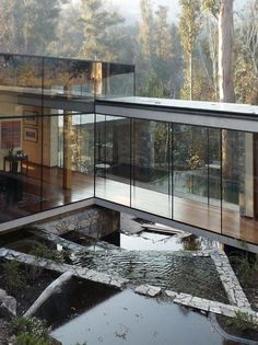 a glass house in the middle of a forest with water running through it and lots of windows