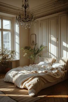 an unmade bed in a room with two windows and a chandelier hanging from the ceiling