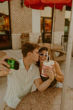 a man and woman kissing while sitting at an outdoor table with drinks in front of them