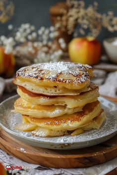 a stack of pancakes covered in powdered sugar on top of a white plate next to apples