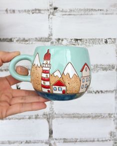 a hand holding a coffee cup with a lighthouse on the side and mountains painted on it