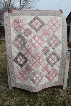 a pink and gray quilt hanging from a clothes line in the grass near a house