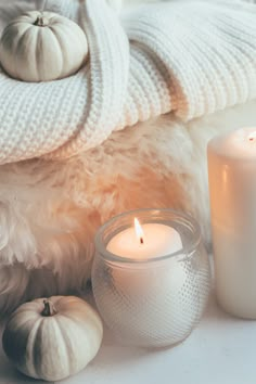 two white candles sitting on top of a table next to some pumpkins and blankets