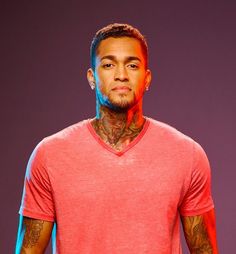 a man with tattoos standing in front of a purple and blue background wearing a red v - neck shirt