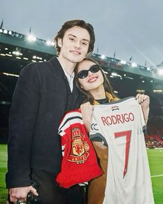 a man and woman posing for a photo in front of a soccer stadium holding a jersey