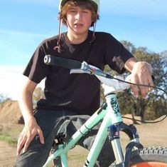 a young man sitting on top of a blue bike next to a dirt field with trees in the background