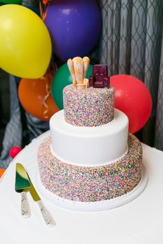 a three tiered cake with colorful sprinkles and balloons in the background