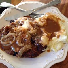 a white plate topped with meat covered in gravy and onions next to mashed potatoes