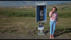 a woman standing next to a phone booth in the middle of a field with mountains behind her