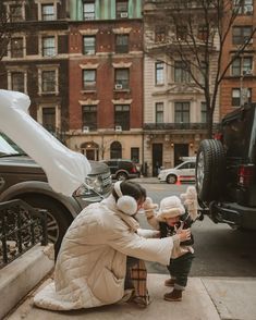 a woman kneeling down next to a child in front of a parked car on the sidewalk