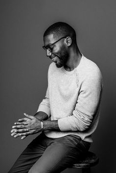 a black and white photo of a man sitting on top of a stool with his hands together