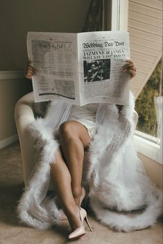 a woman is sitting on a couch reading a newspaper while wearing white fur coat and high heels