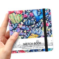 a hand holding a sketch book in front of a white background with blue and pink flowers