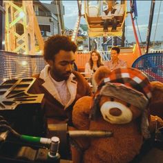 a man sitting next to a brown teddy bear on top of a roller coaster at an amusement park