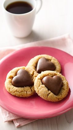 three heart shaped cookies on a pink plate next to a cup of coffee