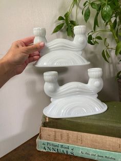 a person is holding a white candle holder on top of a stack of books next to a potted plant