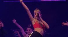 a woman in a red top and black shorts on stage with her arms raised up