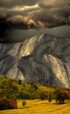 a large mountain covered in lots of clouds next to a lush green field with trees