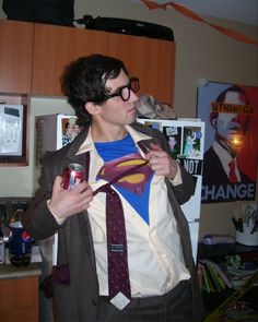 a man wearing a superman t - shirt and tie in a kitchen with posters on the wall