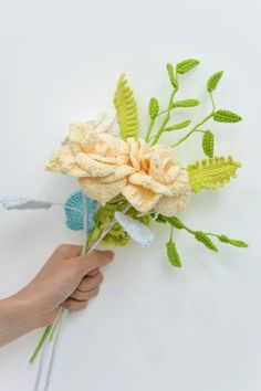 a hand holding a flower made out of crocheted yarn and leaves on a white background