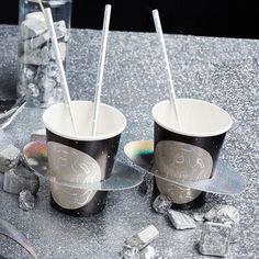 two cups with straws in them sitting on a table next to ice cubes