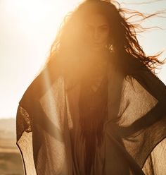 a woman standing in the sun with her hair blowing in the wind and wearing a sheer shirt