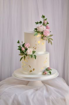 a three tiered cake with pink flowers and greenery on the top is sitting on a white tablecloth