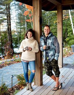 a man and woman standing next to each other on a wooden porch with trees in the background