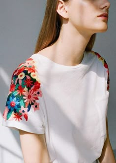 a woman with long hair wearing a white shirt and floral embroidery on her sleeves, looking off to the side