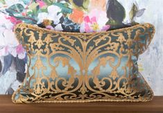 a blue and gold pillow sitting on top of a wooden table next to a floral wallpaper