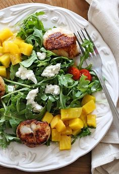 a white plate topped with salad and meat on top of a wooden table next to a fork