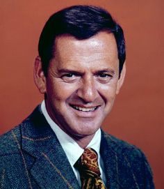 a man in a suit and tie smiling at the camera with an orange background behind him