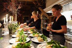 three women preparing food in a kitchen with candles on the counter and flowers behind them