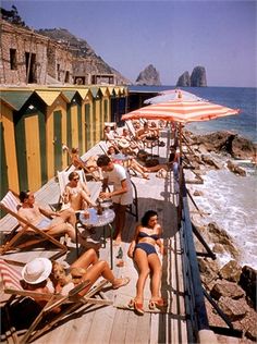 people are lounging on the boardwalk near the water and beach huts with umbrellas