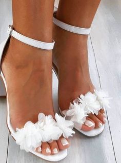 a woman wearing white high heels with flowers on the toe and ankle straps, standing in front of a wooden floor