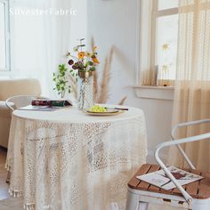 a table with a plate and flowers on it in front of a chair next to a window