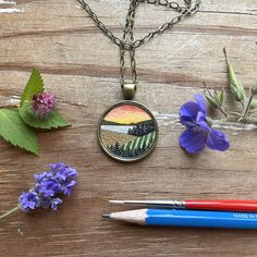 a pen and some purple flowers on a wooden table next to a necklace with an image of a bridge