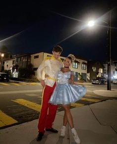 a man and woman dressed up in costumes standing on the sidewalk at night with street lights behind them