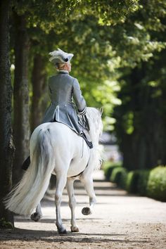 a woman riding on the back of a white horse down a road next to trees