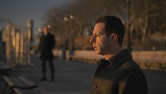 a man standing next to a wooden bench on a city street at night with another man in the background