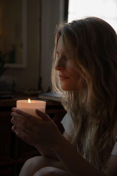 a woman holding a lit candle in her hands while sitting on the floor next to a window