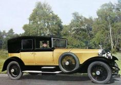 an old yellow car is driving down the road in front of some trees and grass