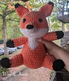 a hand holding an orange and white crocheted stuffed animal in front of trees