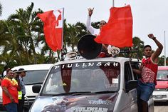 two men waving red flags on top of a car with other people in the background