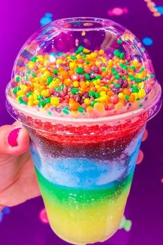 a person holding up a cup filled with rainbow colored liquid and sprinkles